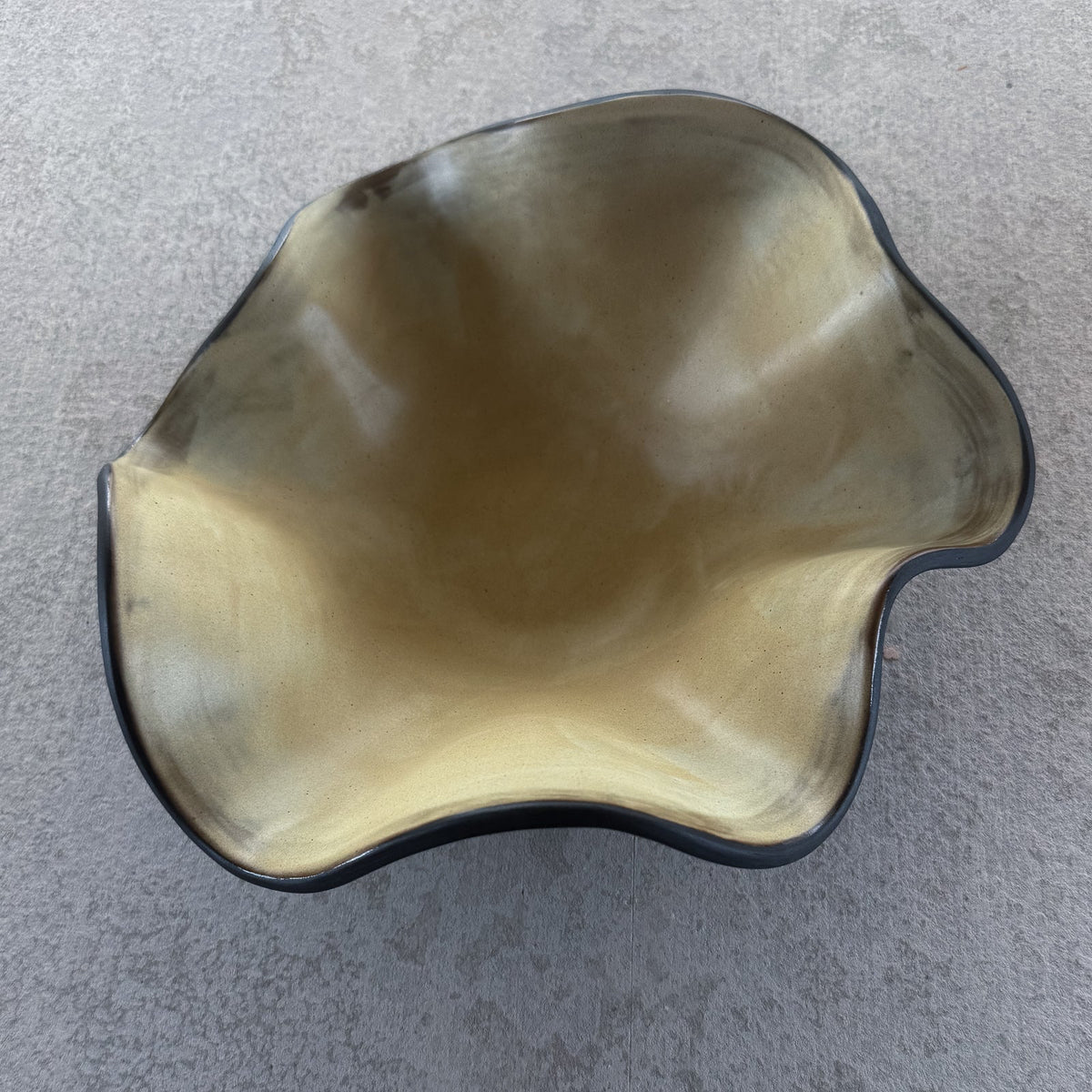 An organic-shaped bowl with a metallic gold interior and a dark charcoal gray exterior, photographed from a top-down perspective on a textured gray concrete surface.