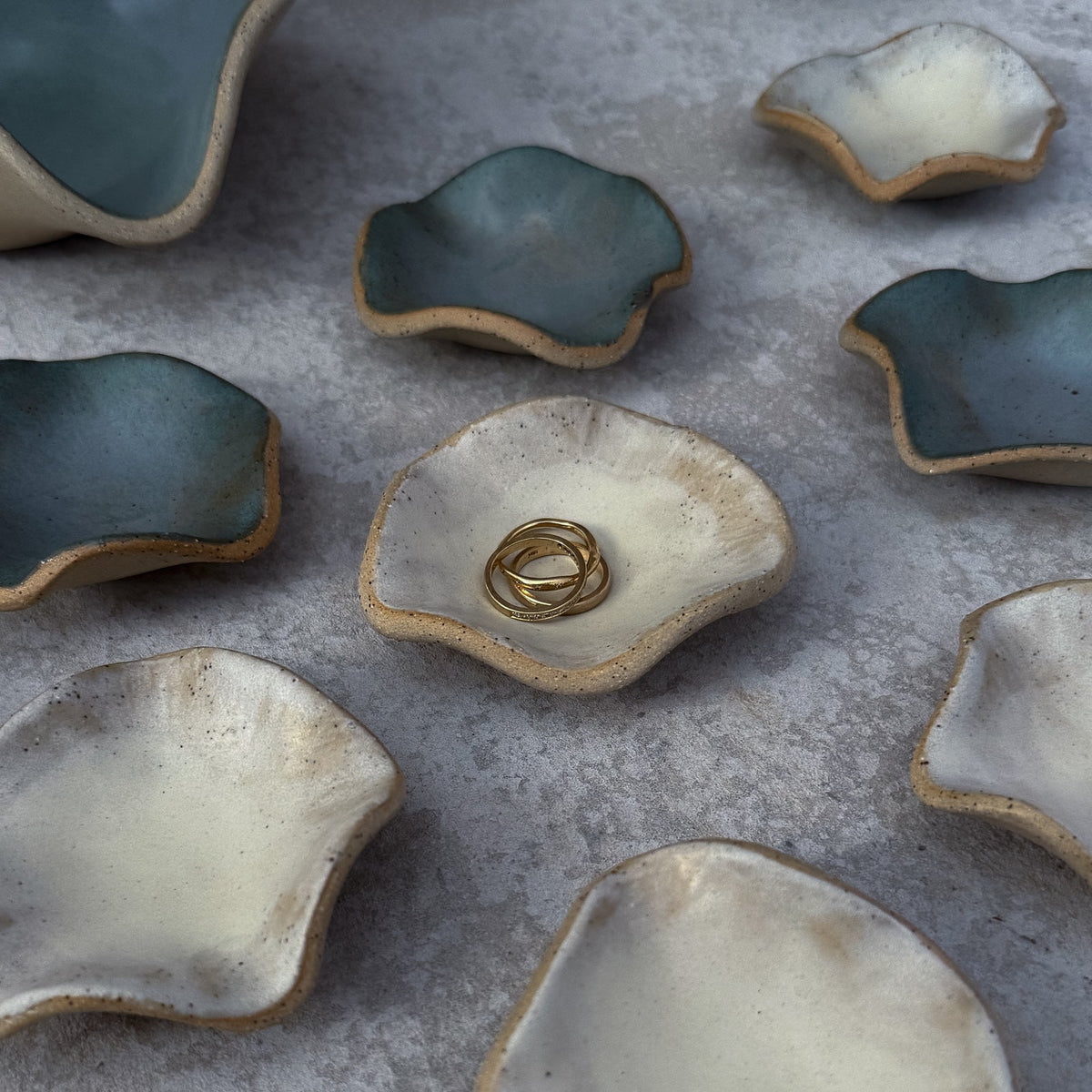 A set of handmade, organic-shaped ceramic ring stands in a flat-lay arrangement on a textured gray surface. The ring stands feature a glossy, mottled glaze of teal-blue and cream-white on the inside, with a rustic, unglazed beige clay edge.