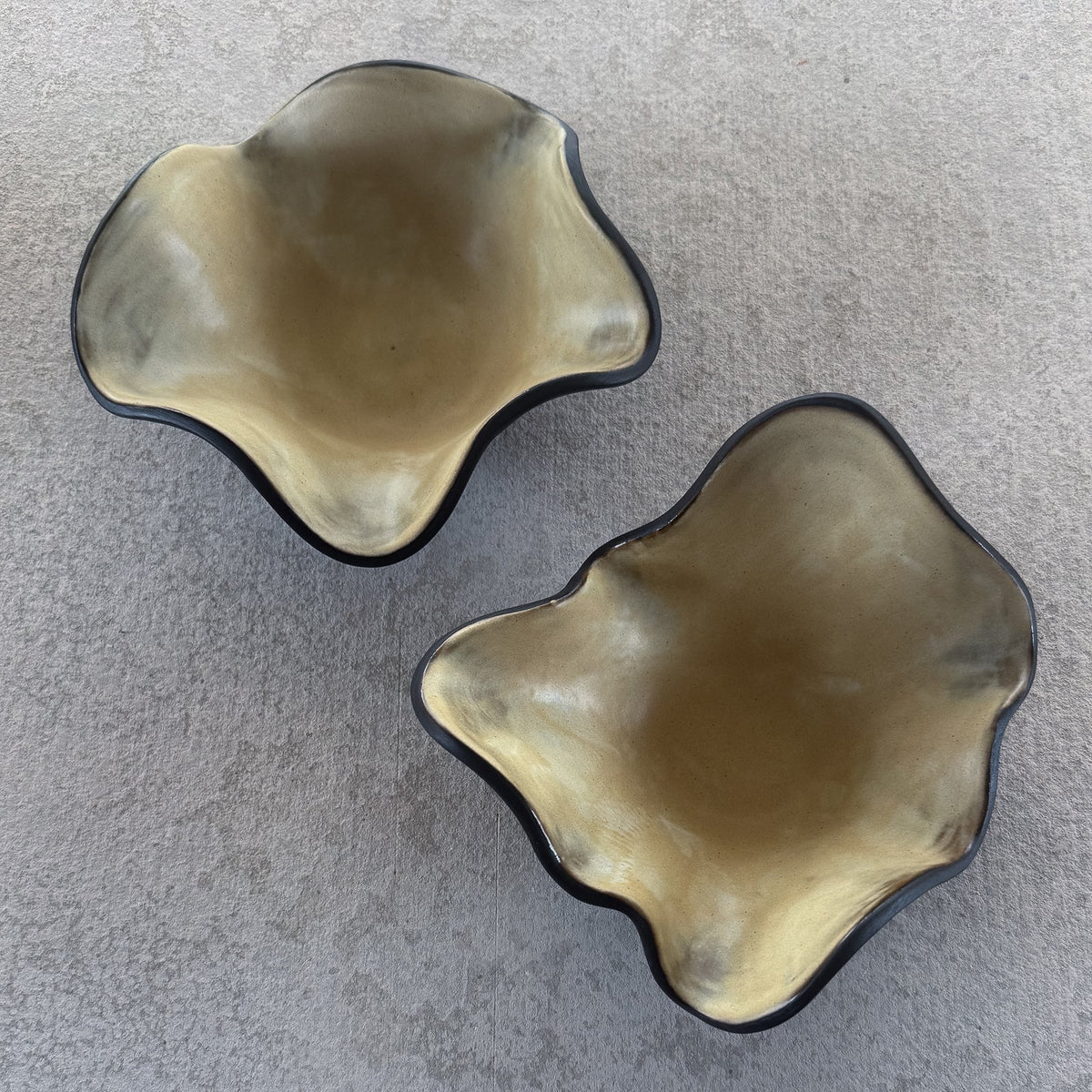 Two organic-shaped ceramic bowls with wavy edges, featuring a glossy metallic gold interior and a dark charcoal gray exterior, displayed on a textured gray concrete surface.